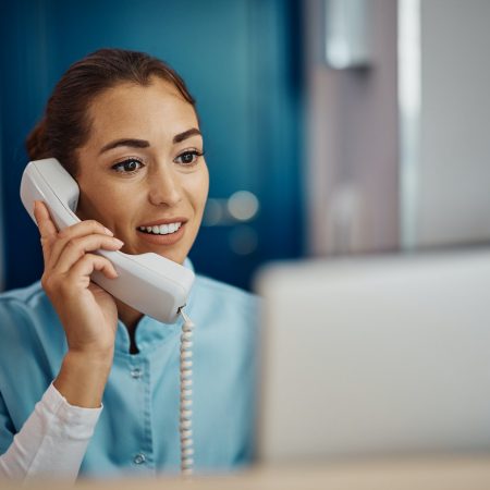 Young nurse answering phone call while working on desktop PC at reception desk in hospital.